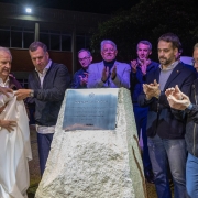Quatro homens revelam uma placa comemorativa em uma pedra rústica, enquanto outras autoridades e convidados aplaudem ao fundo, em evento noturno.