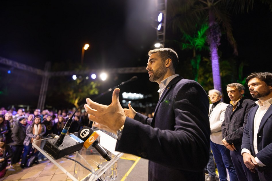 O Governador Eduardo Leite discursa em evento noturno, gesticulando em um pódio transparente com microfones. Uma plateia atenta e outras autoridades o observam.