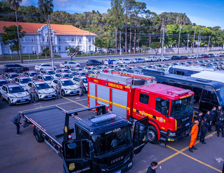 A imagem mostra agentes de segurança pública de pé ao lado de um veículo grande dos bombeiros, que tem detalhes em vermelho, branco e amarelo. O veículo está estacionado em um pátio amplo, sob céu limpo e ensolarado. Ao fundo, há outros veículos e estruturas da área onde ocorre o evento.