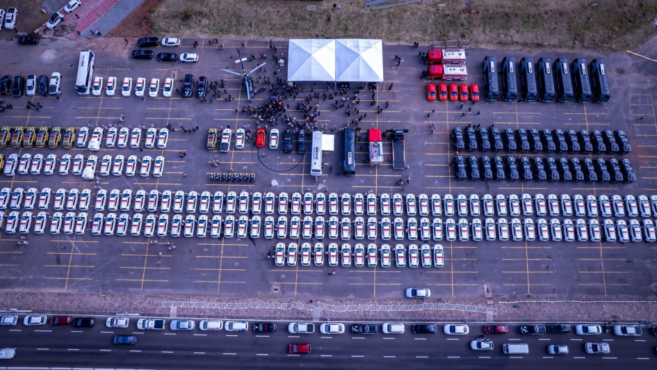 Foto aérea, tirada por drone, que mostra uma grande quantidade de viaturas alinhadas em fileiras simétricas. Há uma concentração de pessoas reunidas em frente a um palco, onde há uma tenda branca. Ao lado do palco, vê-se um helicóptero exposto. Toda a cena acontece em um pátio grande e aberto, cercado por áreas verdes.