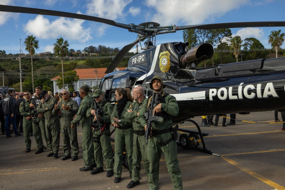A foto mostra um helicóptero preto, pousado no pátio. O helicóptero tem a inscrição da polícia. Agentes de segurança estão alinhados em frente ao veículo.
