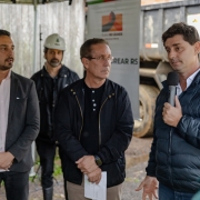 Há 5 pessoas de pé. Quatro estão de frente e um olha para eles. Dois estão de mãos cruzadas e um segura um microfone. Ao fundo há outro homem que usa capacete de obras. Ao fundo das pessoas aparece uma roda de caminhão.