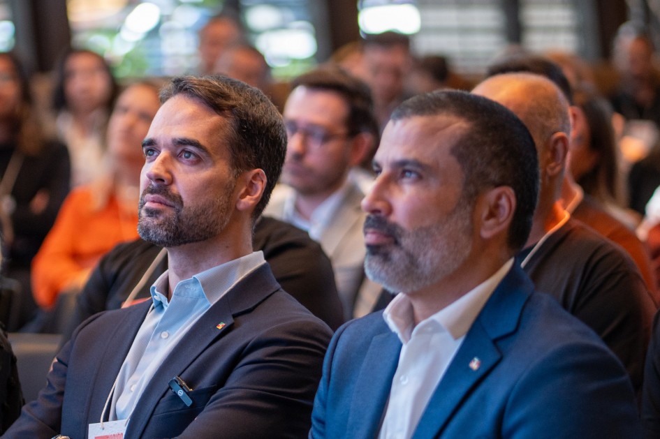 Há duas pessoas com foco na foto e ao fundo diversas outras pessoas. Todos aparecem estar sentados.