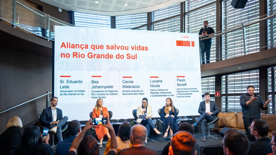 Há seis pessoas no palco. Cinco estão sentadas e uma de pé. Uma mulher no centro do palco está com o microfone na mão. Atrás deles há um painel.