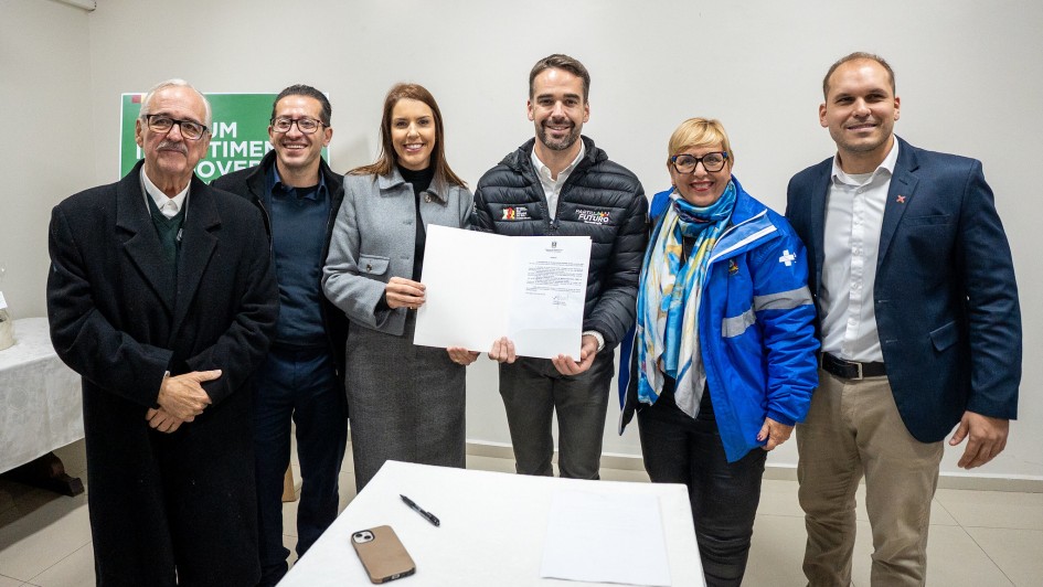 Seis pessoas posam sorrindo, segurando um documento. No centro, o governador com jaqueta preta e a secretária-adjunta de blazer cinza mostram o papel. Ao redor, três homens e uma mulher de jaqueta azul. Na frente, uma mesa com celular, caneta e papel.