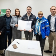 Seis pessoas posam sorrindo, segurando um documento. No centro, o governador com jaqueta preta e a secretária-adjunta de blazer cinza mostram o papel. Ao redor, três homens e uma mulher de jaqueta azul. Na frente, uma mesa com celular, caneta e papel.