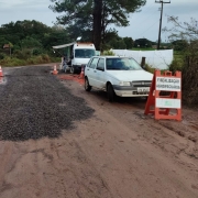 A imagem mostra uma estrada de terra com alguns trechos cobertos por cascalho. No lado direito, há um carro branco parado ao lado de um cavalete laranja com uma placa que diz “FISCALIZAÇÃO AGROPECUÁRIA”. Atrás do carro, há um furgão branco com um toldo estendido, sob o qual estão pessoas sentadas em cadeiras, aparentemente responsáveis pela fiscalização. Cones de sinalização estão espalhados pela via, delimitando o local da abordagem. O cenário é rural, com vegetação, árvores e uma pequena construção ao fundo. O dia está nublado e o chão está molhado, com poças de água.