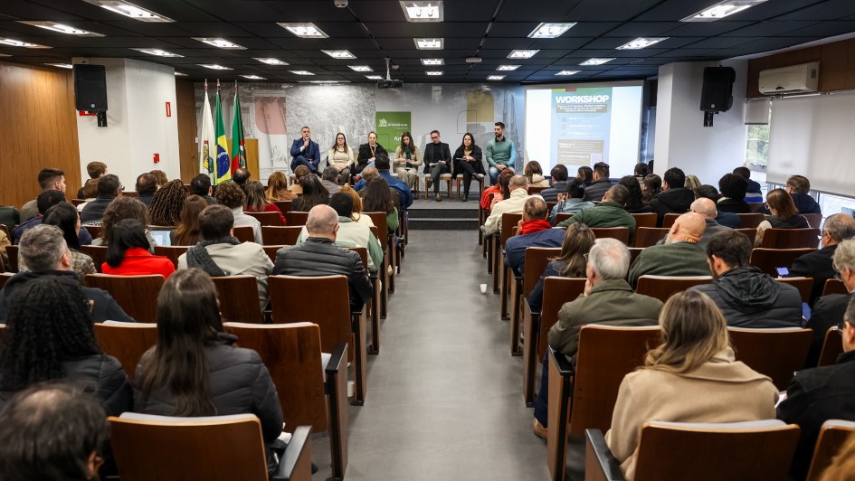 Em uma sala há dezenas de pessoas sentas olhando para o palco. No palco há sete pessoas sentadas. O homem da esquerda está com o microfone na mão. Tem três bandeiras ao lado, uma do Estado do Rio Grande do Sul, outra do Brasil e outra branca. Do lado direito tem um painel de apresentação.