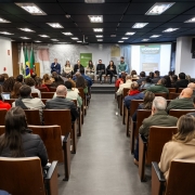 Em uma sala há dezenas de pessoas sentas olhando para o palco. No palco há sete pessoas sentadas. O homem da esquerda está com o microfone na mão. Tem três bandeiras ao lado, uma do Estado do Rio Grande do Sul, outra do Brasil e outra branca. Do lado direito tem um painel de apresentação.