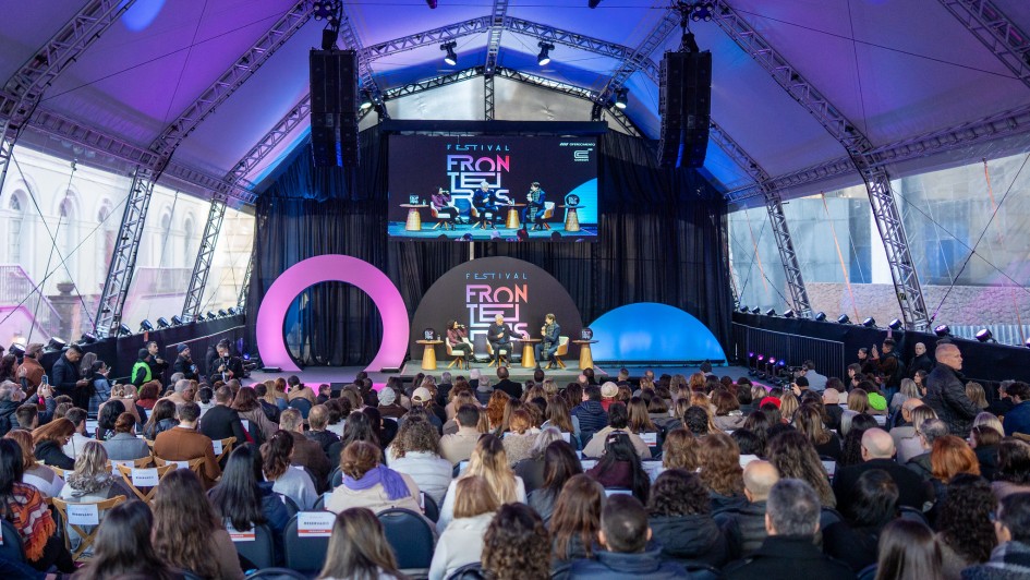 Há um palco colorido com uma apresentação com pessoas sentadas dialogando e uma plateia assistindo.