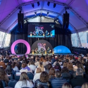 Há um palco colorido com uma apresentação com pessoas sentadas dialogando e uma plateia assistindo.