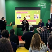 Foto de um homem em pé falando ao microfone. Outros dois painelistas estão sentados observando. Parte da plateia pode ser vista de costas, em Painel sobre Centro Agro na Gramado Summit 2025