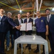 Na foto, sete homens estão em pé, sorrindo, posando para foto. Ao centro, Leite segura um documento aberto, visível para a câmera, enquanto os outros encostam as mãos no papel ou no ombro uns dos outros. Eles estão em um espaço interno simples, com teto de madeira e fios amarelos cruzando no alto. À frente deles há uma mesa branca de plástico, vazia, exceto por uma caneta. Ao fundo, outras pessoas observam a cena.