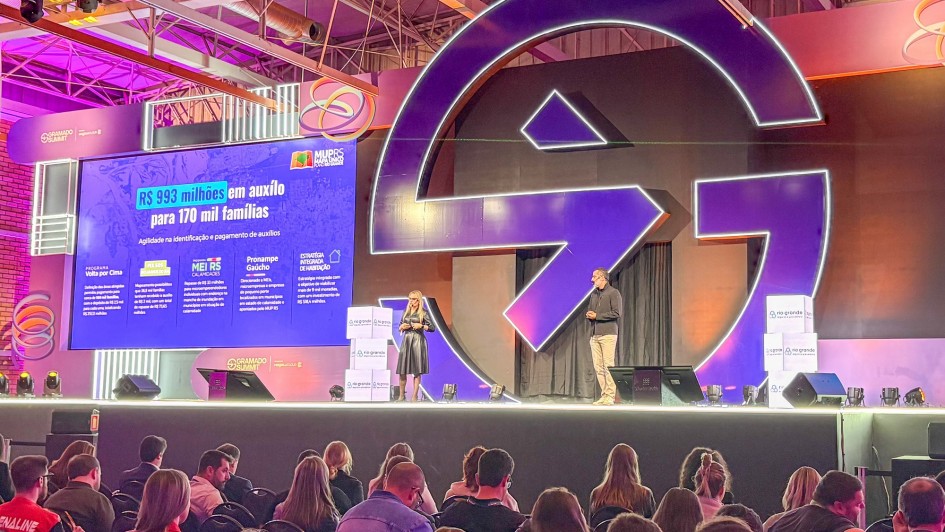Danielle Calazans e Bruno Silveira em pé sobre o palco principal do evento fazendo a apresentação na abertura do Gramado Summit 2025. Parte da plateia pode ser vista de costas.