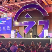 Danielle Calazans e Bruno Silveira em pé sobre o palco principal do evento fazendo a apresentação na abertura do Gramado Summit 2025. Parte da plateia pode ser vista de costas.