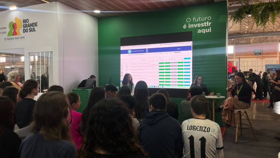 H&aacute; dezenas de crian&ccedil;as de costas, sentadas, assistindo uma apresenta&ccedil;&atilde;o. No palco h&aacute; quatro pessoas. Dias mulheres fazem a apresenta&ccedil;&atilde;o, enquanto uma menina assiste e um homem est&aacute; sentado de frente ao um notebook. H&aacute; um painel com informa&ccedil;&otilde;es atr&aacute;s deles.