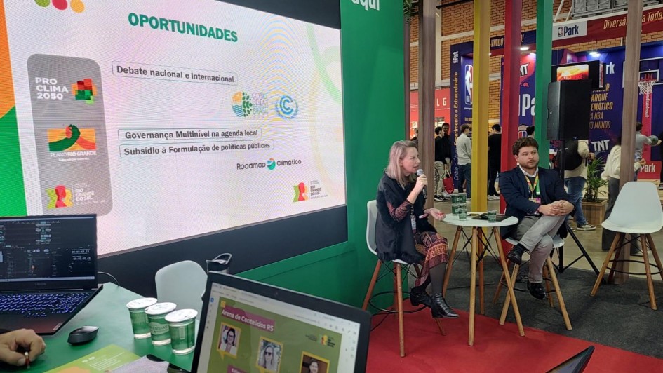 Há duas pessoas sentadas e uma mesa pequena redonda no centro entre elas. A mulher segura um microfone. O homem está de pernas cruzadas escutando. Há dois computadores sobre uma outra mesa. Ao fundo há um painel de apresentação.