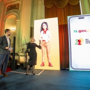 Callazans e Leite em pé sobre um palco apresentando a GurIA, inteligência artificial do rs.gov.br. Há dois cartazes grandes um deles mostrando a imagem do avatar da nova IA do Estado.