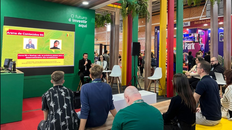 Foto mostra um evento de tecnologia e inova&ccedil;&atilde;o em ambiente interno moderno, com destaque para uma apresenta&ccedil;&atilde;o no "Arena de Conte&uacute;dos RS" sobre "Inova RS e Startup Lab". Dois palestrantes - um homem de terno com microfone e uma mulher - apresentam para uma plateia de cerca de 20 pessoas sentadas em cadeiras brancas e amarelas, em um espa&ccedil;o decorado com pilares coloridos (verde, amarelo e vermelho), teto de madeira, plantas suspensas e outros estandes ao fundo, criando uma atmosfera contempor&acirc;nea de networking e empreendedorismo no Rio Grande do Sul.