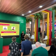 Foto mostra um evento de tecnologia e inovação em ambiente interno moderno, com destaque para uma apresentação no "Arena de Conteúdos RS" sobre "Inova RS e Startup Lab". Dois palestrantes - um homem de terno com microfone e uma mulher - apresentam para uma plateia de cerca de 20 pessoas sentadas em cadeiras brancas e amarelas, em um espaço decorado com pilares coloridos (verde, amarelo e vermelho), teto de madeira, plantas suspensas e outros estandes ao fundo, criando uma atmosfera contemporânea de networking e empreendedorismo no Rio Grande do Sul.