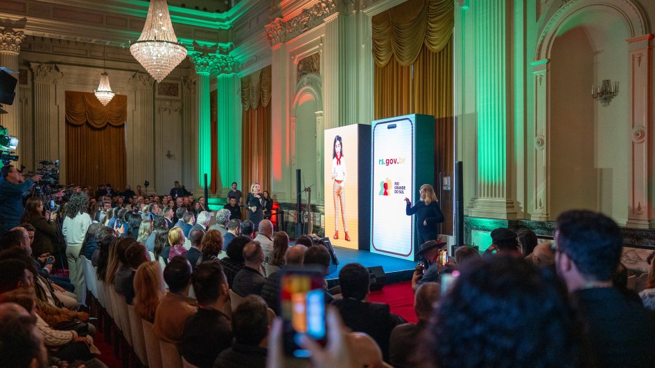 Dezenas de pessoas assistem a uma palestra. Há uma tela em formato de celular gigante com imagens aparecendo. Há ma mulher d epé com o microfone na mão.