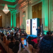 Dezenas de pessoas assistem a uma palestra. Há uma tela em formato de celular gigante com imagens aparecendo. Há ma mulher d epé com o microfone na mão.