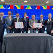 Há sete pessoas de pé posando para foto. Uma mesa com pano cinza escuro está à frente deles. Ao fundo um painel azul e com cores que lembram as bandeiras do Brasil e da França. Três seguram dois documentos abertos e assinados.