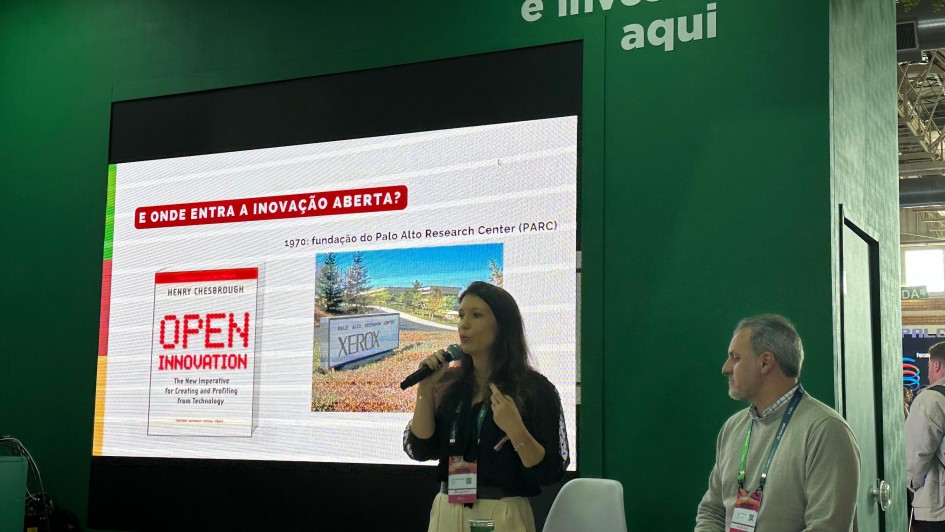 Há duas pessoas em um palco. Uma mulher segura um microfone na mão. Ela está de pé e parece falar. Um homem está sentado, e olha para ela. Ao fundo há um painel de apresentação em uma parede verde.