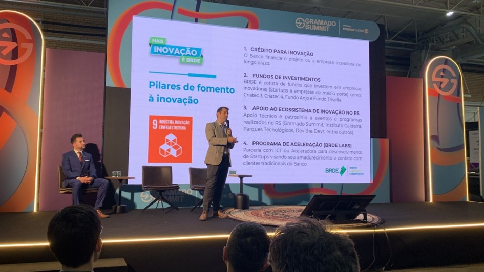Imagem de duas pessoas em um palco durante painel do BRDE no Gramado Summit 2025. Uma delas está em pé e com o microfone na mão falando.