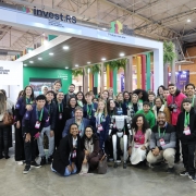 Foto de um grande grupo de pessoas reunidas em frente ao estande do Governo do Rio Grande do Sul, em um evento realizado em um pavilhão coberto. A maioria usa crachás coloridos e colares verdes. Ao centro, há um robô humanoide com braços estendidos, rodeado por pessoas sorridentes, muitas delas jovens. O ambiente tem decoração moderna, com cores vivas, plantas e letreiros.
