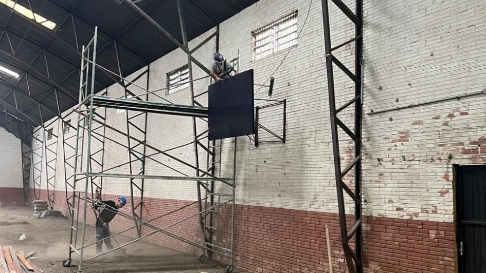 Imagem mostra reforma de ginásio do Colégio Protásio Alves. Homem trabalha em andaime instando tabela de basquete. Outro homem varre o chão do ginásio