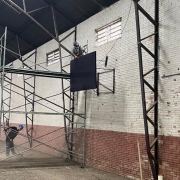 Imagem mostra reforma de ginásio do Colégio Protásio Alves. Homem trabalha em andaime instando tabela de basquete. Outro homem varre o chão do ginásio