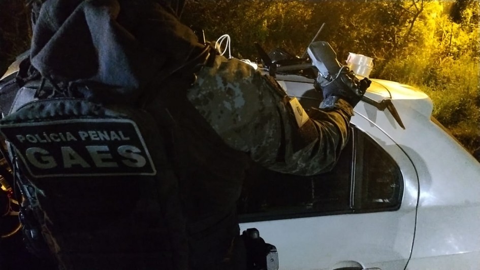 Imagem mostra policial com coleta da Polícia Penal Gaes segurando drone com a mão direita, sobre um veículo de cor branco