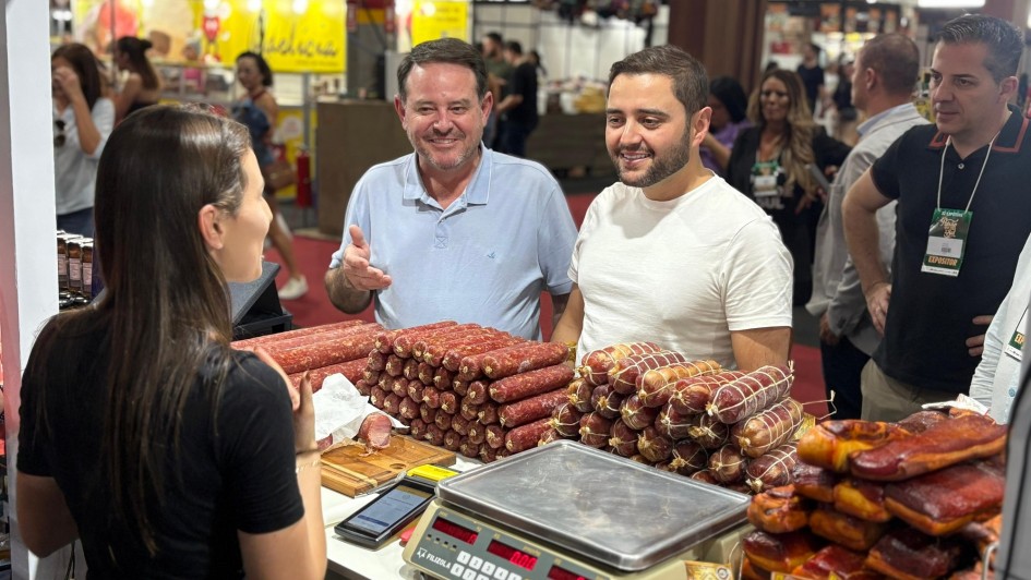 Há três homens de pé, de frente para uma mulher que atende eles. Na bancada há diversos tipos de comida do tipo embutidos.