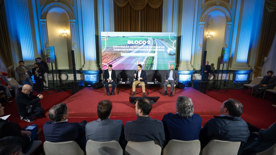 Leite ao microfone no anúncio. Ele e dois outros homens estão sentados diante de uma tela e parte da plateia pode ser vista de costas no anúncio de mudanças na concessão do Bloco em 2025.