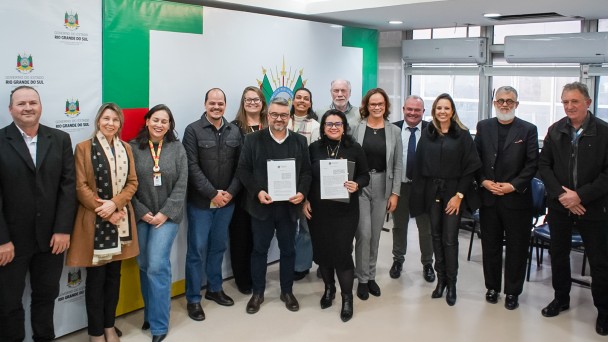 Foto posada de diversas pessoas reunidas em torno de duas delas que seguram os documentos recém assinados.