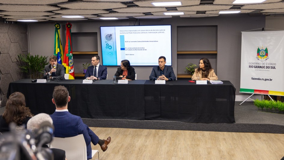 Foto da mesa com cinco pessoas concedendo entrevista coletiva sobre descontos em folha de pagamento de servidores inativos. Duas pessoas da plateia podem ser vistas de costas.