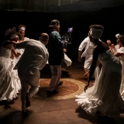 Imagem mostra grupo de atores dançando ao redor de cinegrafista. A luz está focada no meio do grupo, que usa roupas brancas