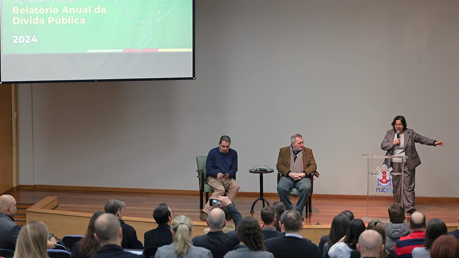 A imagem mostra uma apresentação intitulada "Relatório Anual da Dívida Pública 2024". Há três pessoas no palco: duas estão sentadas em cadeiras e uma está de pé, aparentemente falando ao público. A pessoa de pé está apontando para a tela de projeção. O público está sentado em cadeiras, assistindo à apresentação.