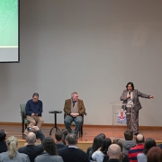 A imagem mostra uma apresentação intitulada "Relatório Anual da Dívida Pública 2024". Há três pessoas no palco: duas estão sentadas em cadeiras e uma está de pé, aparentemente falando ao público. A pessoa de pé está apontando para a tela de projeção. O público está sentado em cadeiras, assistindo à apresentação.