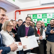 A imagem retrata um evento público em um ambiente interno, onde um grupo de pessoas está reunido, a maioria sorrindo e olhando para o centro da cena.

Em destaque no centro, um homem com barba e blazer escuro, parecendo ser uma figura de autoridade, segura um documento branco, que aparenta ser um certificado ou contrato, e sorri para a câmera. Ele está cercado por outras pessoas.

À esquerda do homem central, uma mulher de cabelos claros e óculos, vestindo um blazer branco, também sorri e parece interagir com o documento ou com o homem ao seu lado. Ao lado dela, parcialmente visível, um homem de camisa escura sorri.

À direita do homem central, um homem mais velho com um chapéu e um lenço vermelho no pescoço está aplaudindo. Atrás deles, há várias outras pessoas, a maioria homens, observando e alguns também aplaudindo.

O fundo da imagem mostra uma parede verde com logotipos repetidos, que parecem ser o nome "BOM PASTOR" e um símbolo de uma cruz, sugerindo o nome de uma instituição. Há também tubulações vermelhas visíveis e um aparelho de ar condicionado no alto da parede. A iluminação é clara e uniforme, indicando um evento bem iluminado. A atmosfera geral é de celebração.