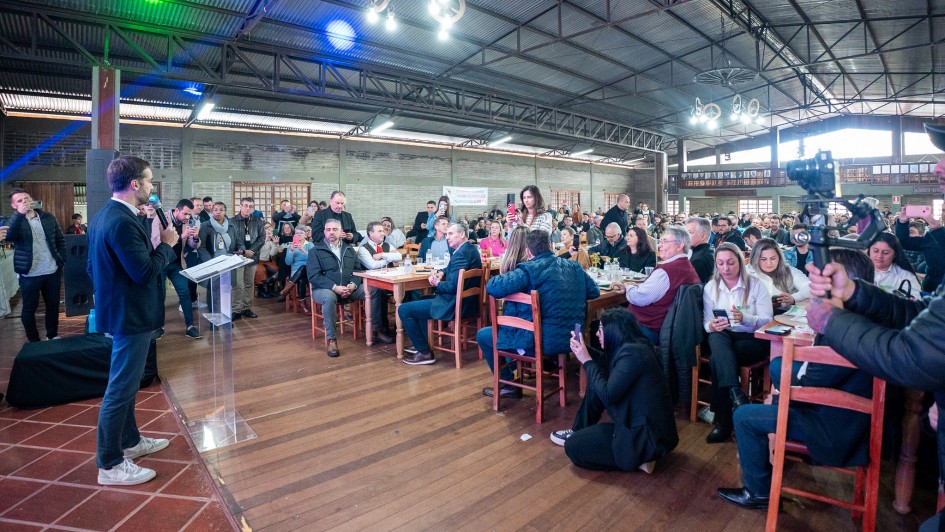 Foto em que Leite aparece à esquerda falando ao microfone, de pé, num ambiente fechado. À direita, a plateia está distribuída em muitas mesas.