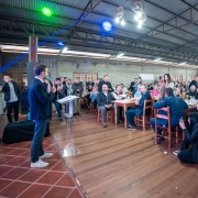 Foto em que Leite aparece à esquerda falando ao microfone, de pé, num ambiente fechado. À direita, a plateia está distribuída em muitas mesas.