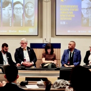 Foto de cinco pessoas estão sentadas à frente de uma plateia, participando de um painel de debate em uma sala de conferência. Atrás deles, um telão exibe os rostos e nomes dos palestrantes, com o título do evento: “Cidades resilientes e inclusivas: desafios e caminhos para infraestruturas sustentáveis”. O ambiente é formal, com todos os participantes vestidos de forma profissional.