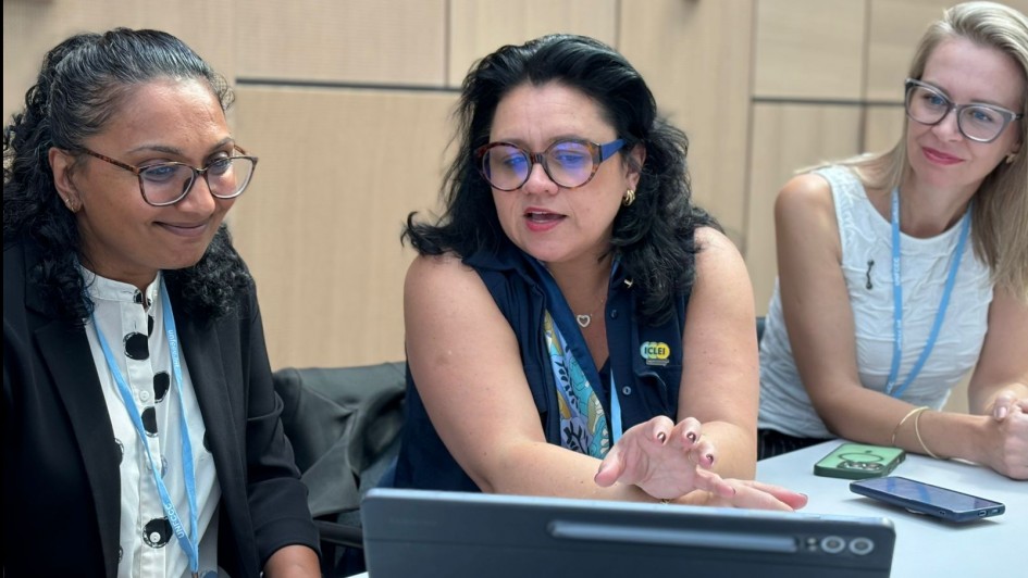 A imagem mostra três mulheres sentadas em uma mesa, aparentemente em uma reunião ou conferência.

A mulher à esquerda tem cabelo escuro e encaracolado e usa óculos. Ela está vestindo um blazer preto sobre uma blusa branca estampada e tem um crachá azul pendurado no pescoço. Ela está olhando para algo à frente com um leve sorriso.

A mulher ao centro tem cabelo escuro e usa óculos grandes e azuis. Ela está vestindo uma blusa azul-marinho e tem um crachá amarelo com a sigla "ICLEI" visível. Ela está gesticulando com a mão direita e parece estar falando ou explicando algo, olhando para uma tela de tablet à sua frente.

A mulher à direita tem cabelo loiro e usa óculos de armação clara. Ela está vestindo uma blusa branca e tem um crachá azul. Ela está olhando para a mulher do centro com atenção e um leve sorriso.

Sobre a mesa, há um tablet preto aberto, que parece ser o foco da atenção da mulher do centro. Também é possível ver um ou dois smartphones sobre a mesa. O ambiente parece ser uma sala de conferências com paredes claras.