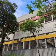 Esta é a fachada da Escola Estadual de Ensino Fundamental Antônio Vieira, localizada em Novo Hamburgo. O prédio tem dois andares, várias janelas e unidades de ar-condicionado instaladas. Ele é cercado por uma grade metálica e há vegetação ao redor. A parte superior do edifício tem uma faixa vermelha, enquanto a parte inferior exibe uma faixa amarela. O céu está azul com algumas nuvens, trazendo uma atmosfera agradável. Parece ser um ambiente escolar bem estruturado!