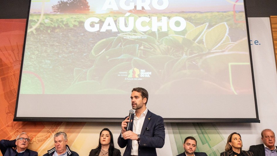 A imagem mostra um evento relacionado ao agro gaúcho, realizado em 16 de outubro de 2023. No palco, há um grande telão exibindo esse título, acompanhado de uma imagem agrícola com o sol ao fundo. Abaixo da tela, seis pessoas estão sentadas em uma mesa com microfones e garrafas de água, enquanto uma pessoa está em pé, falando ao público. O ambiente sugere uma conferência ou reunião focada em discussões sobre agricultura no Rio Grande do Sul. Parece ser um evento importante para debater desafios e oportunidades do setor.