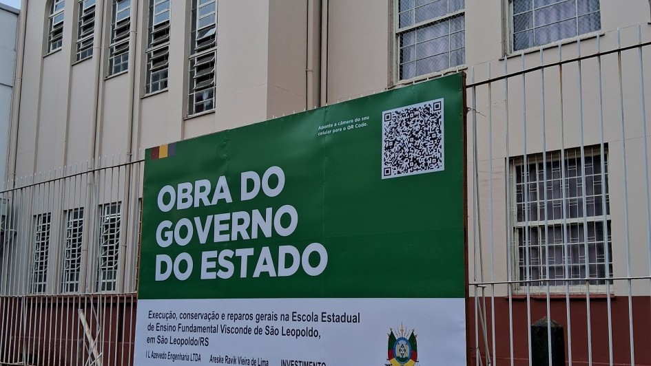 A imagem mostra um prédio com várias janelas e um cartaz verde e branco preso a uma cerca na frente do edifício. O cartaz contém texto em português, um QR code e informações sobre um projeto governamental de manutenção e reparos na Escola Estadual de Ensino Fundamental Visconde de São Leopoldo, localizada em São Leopoldo, Rio Grande do Sul, Brasil. Também há detalhes sobre a empresa responsável pela obra e o investimento total, que é de R$ 1.287.670,11. O QR code convida os espectadores a escaneá-lo para obter mais informações. A cena destaca um investimento público na infraestrutura escolar da região.