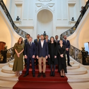 A imagem mostra um grupo de pessoas em uma escadaria grandiosa dentro de um edifício elegante. A arquitetura apresenta detalhes ornamentados, esculturas e luminárias decorativas. Os presentes estão vestidos formalmente, indicando um evento especial ou cerimônia oficial.