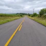 A imagem mostra uma estrada asfaltada de pista única que se estende ao longe sob um céu nublado. A estrada é cinza-escura e apresenta marcações amarelas no centro: uma linha contínua no lado esquerdo e uma linha tracejada no lado direito, indicando as faixas de tráfego e regras de ultrapassagem. Em ambos os lados da estrada, há faixas de grama verde vibrante e vegetação rasteira, com algumas árvores e arbustos mais distantes. No horizonte, pode-se ver morros e mais vegetação. À direita da estrada, um poste de madeira com fios elétricos se destaca contra o céu. A cena transmite uma sensação de tranquilidade e natureza, com a estrada convidando a uma jornada.